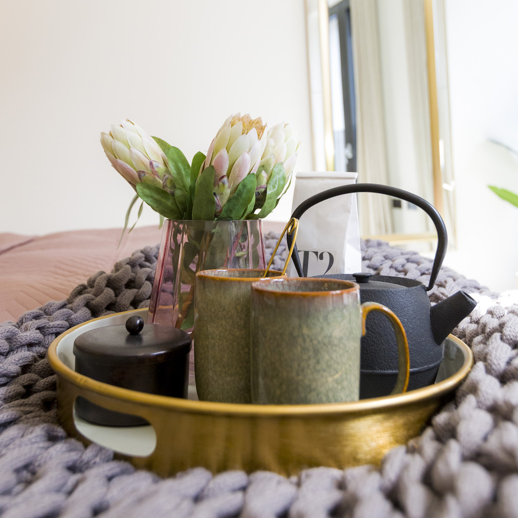 Close up of tea pot, cups and flowers in a vase, place on the a blanket at the bottom of double bed