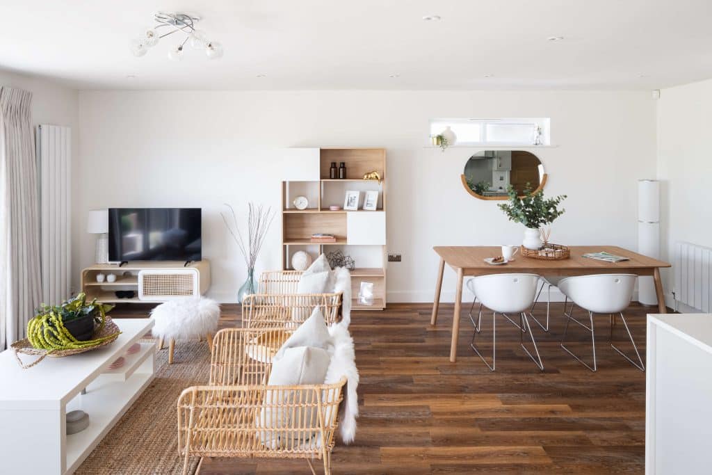 Open space of living room and dining room, with dark laminated flooring, rattan furniture, and white accessories of varied textures