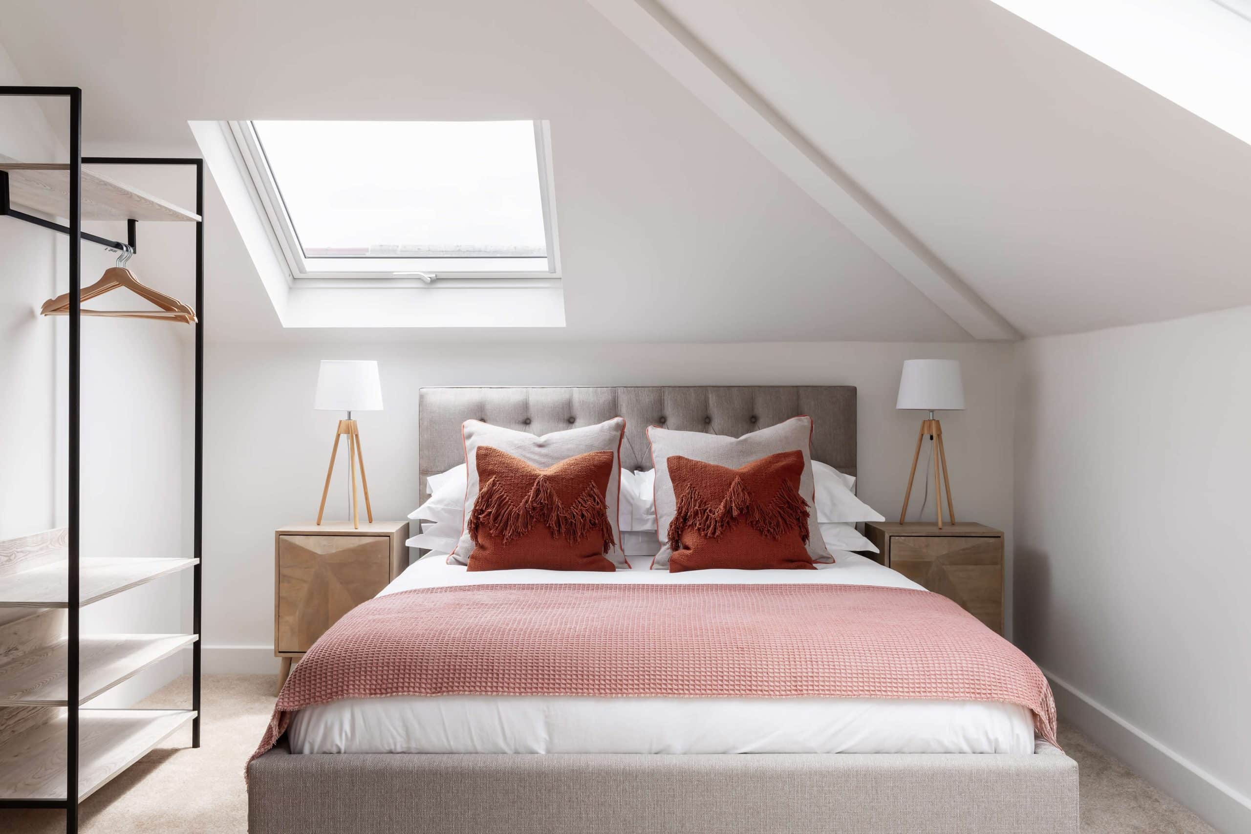 Master bedroom of muted warm tones of dark pink on a light grey bed. Surrounded with symmetrical nightstands and lamps with an empty open wardrobe on the left, under a sunroof window