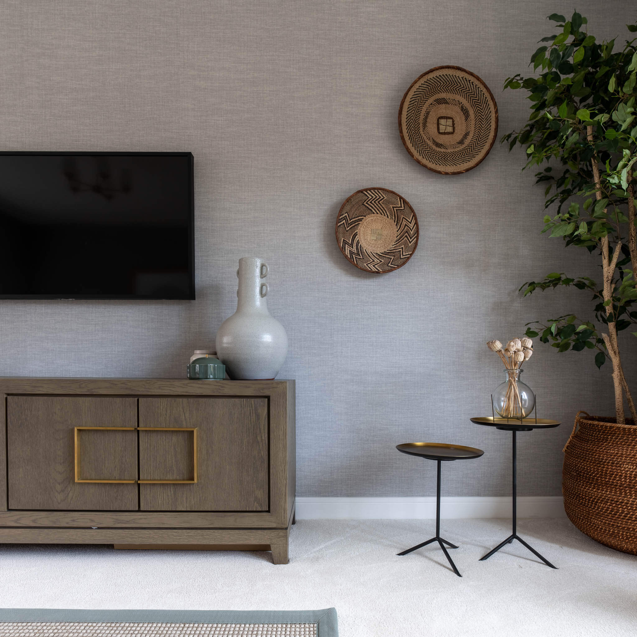 A picture of a neutral coloured living room, with a TV mounted above a console on the left, and small tables, accessories and a plant are on the right